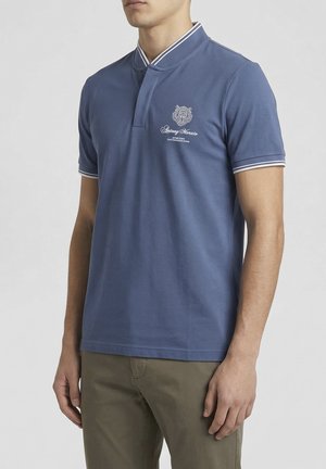 Man wearing blue short-sleeve polo shirt with white striped collar and cuffs, embroidered logo on chest, paired with olive green pants.