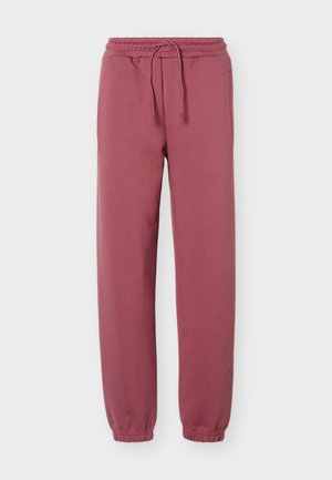 Personne debout portant un haut gris ajusté à manches longues avec fermeture éclair, un pantalon rose ample à cordon et des baskets blanches sur fond blanc.