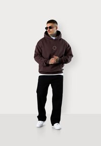Sweatshirt à capuche marron avec un logo, porté sur un T-shirt blanc, associé à un pantalon noir et des baskets blanches. Des lunettes ajoutent une touche élégante.