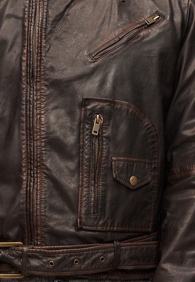 Κοντινό πλάνο του μπροστινού μέρους σκούρας καφέ δερμάτινου μπουφάν που δείχνει κάθετες και διαγώνιες τσέπες με φερμουάρ, τσέπη με καπάκι και κουμπί σνάπ, και ζώνη με μπρούτζινη πόρπη.