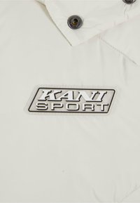 Bijela puffer jakna s istaknutim crnim i srebrnim logotipom "KANI SPORT". Uključuje srebrne gumbe na drukere i kviltanu teksturu.
