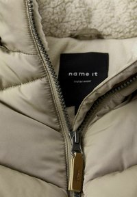Bežs polsterēts džemperis ar sudraba rāvējslēdzēju, ar mīkstu flīsa oderi un melnu birku, uz kuras rakstīts "name it outerwear."