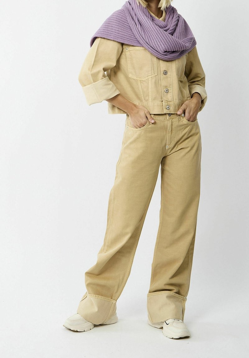 Personne portant un pantalon large beige et une veste courte assortie, avec une écharpe en tricot violet drapée sur les épaules et des baskets blanches.