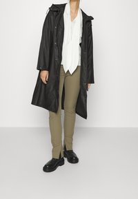 Abrigo largo negro impermeable con cordones, llevado sobre una camisa blanca y pantalones caqui, con dobladillos con aberturas y zapatos gruesos negros.