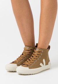 Wysokie sneakersy z brązowego płótna z białym noskiem i podeszwą. Mają beżowe sznurówki oraz charakterystyczne białe logo na boku.