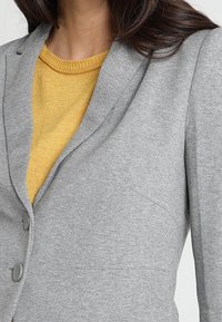 Blazer gris con dos botones, hecho de tela texturizada. Presenta un solapa puntiaguda y un bolsillo lateral, llevado sobre un suéter amarillo.