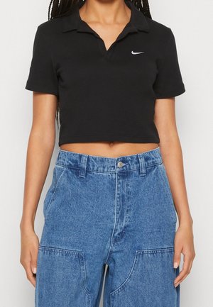 Femme portant un polo noir court avec un petit logo Nike blanc et un jean bleu taille haute avec de grandes poches avant.