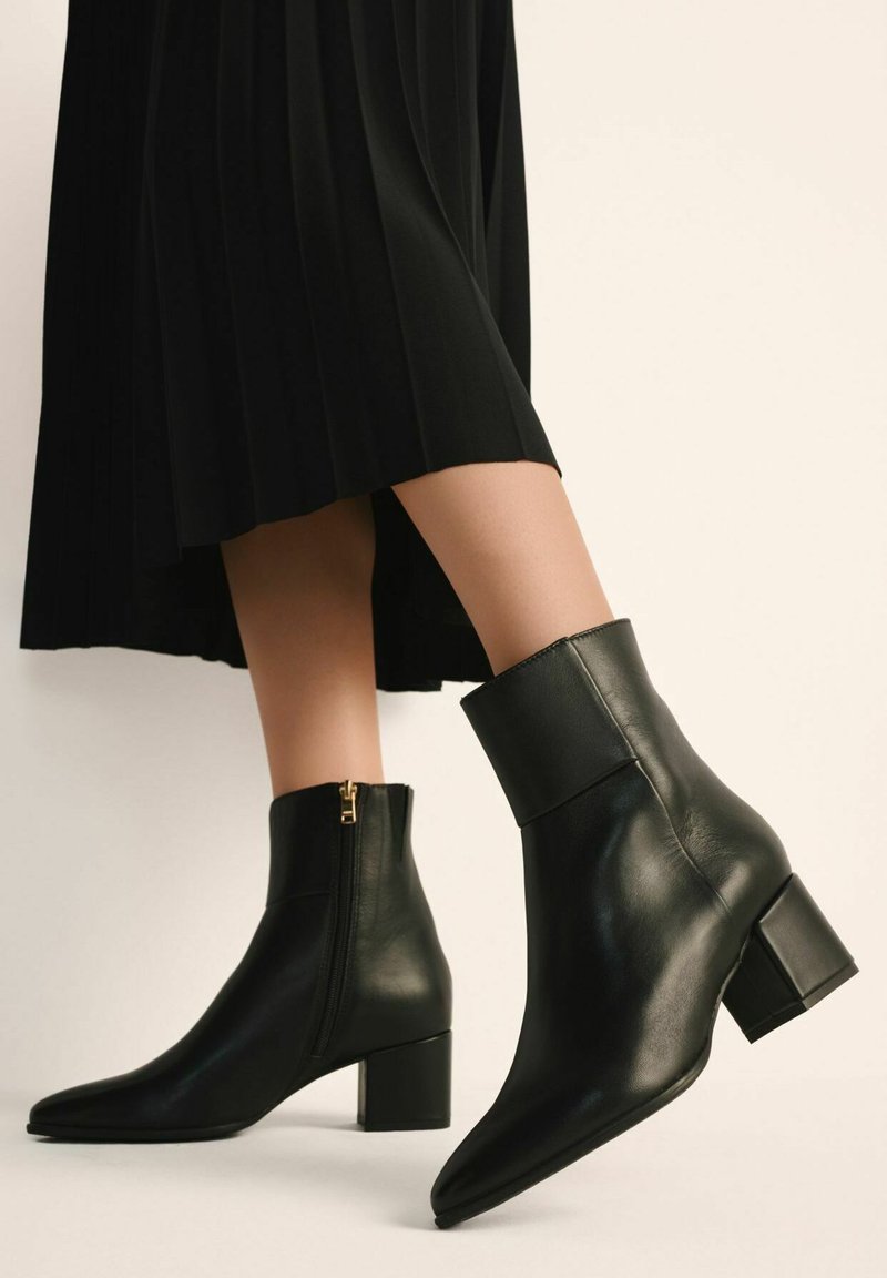 Czarne skórzane botki za kostkę z wąskim noskiem, szerokim obcasem i bocznym zamkiem, z złotym akcentem. Noszone z plisowaną czarną spódnicą.