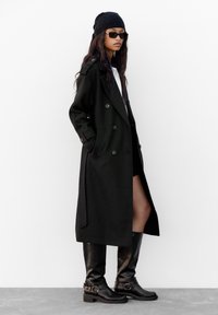 Crni dvoredni kaput s bočnim džepovima, crne kožne čizme do koljena s detaljima kopče i tamna kapa, u kombinaciji s bijelom košuljom.