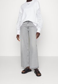 Vit sweatshirt med broderad håldetalj på ärmarna, kombinerad med grå vida jeans och svarta sandaletter med remmar.