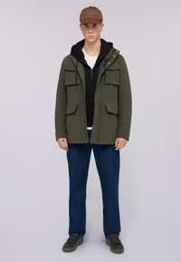 Chaqueta verde oliva con múltiples bolsillos frontales, superpuesta a una sudadera negra con capucha, combinada con jeans azules y zapatos oscuros. Una gorra marrón completa el look.