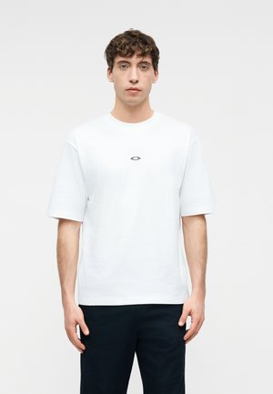 Young man with curly hair stands facing forward wearing a plain white short-sleeve t-shirt with a small black logo on the chest.