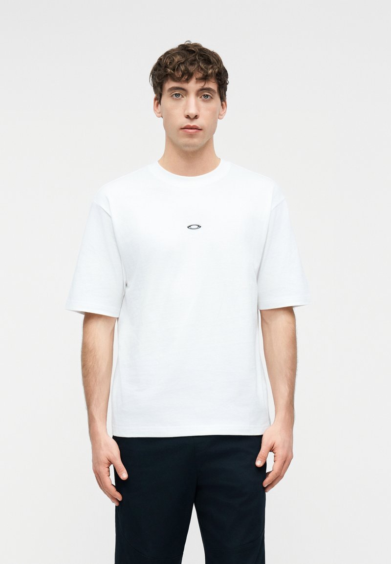 Νεαρός άνδρας με σγουρά μαλλιά στέκεται με το πρόσωπο προς τα εμπρός, φορώντας ένα απλό λευκό κοντομάνικο μπλουζάκι με ένα μικρό μαύρο λογότυπο στο στήθος.