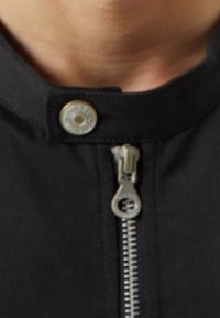 Zwarte jack met een zilveren rits en drukknoopsluiting. Beschikt over een textuurovervoering en zichtbare stiksels langs de kraag en voorkant.