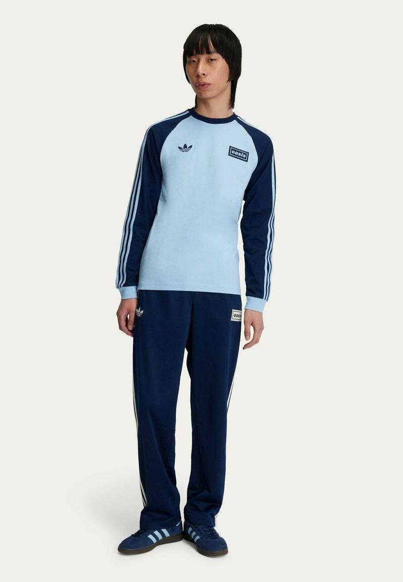 Camicia a maniche lunghe azzurro chiaro con maniche blu navy, tre strisce bianche e un'etichetta "oasis"; abbinata a pantaloni blu navy scuri e scarpe blu navy.