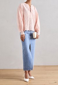 Personne portant un chemisier en dentelle rose, une jupe en denim bleu clair taille haute avec une fente à l'avant, des talons blancs pointus, et tenant une pochette beige.