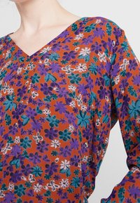 Blusa floreale arancione a scollo a V con fiori viola e teal. Tessuto leggero con una finitura texture. Presenta un sottile dettaglio della tasca frontale.