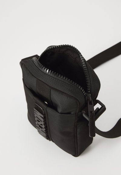Svart tyg crossbody-väska med dragkedjestängning, texturerad yta, platt frontficka och justerbar axelrem. Har en framträdande logotypdetalj.