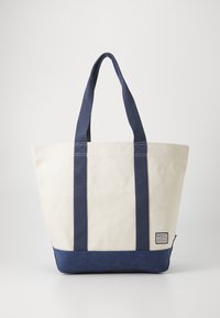 Sac fourre-tout en toile de couleur crème avec des accents bleu marine, doté de poignées robustes et d'un fond solide, avec une étiquette de marque sur le côté.