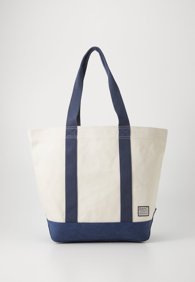 Sac fourre-tout en toile de couleur crème avec des accents bleu marine, doté de poignées robustes et d'un fond solide, avec une étiquette de marque sur le côté.