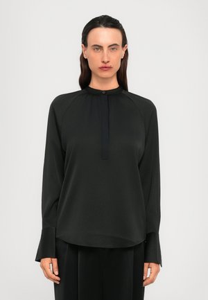 Person with long dark hair wearing a black long-sleeve blouse with a buttoned collar and wide cuffs, standing against a plain background.