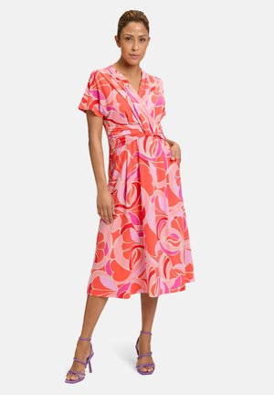 Robe portefeuille à imprimé floral rose avec des manches courtes, un lien à la taille et des poches. Buste ajusté et jupe évasée. Portée avec des talons violets à lanières.
