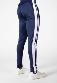 Marineblauwe sportbroek met witte zijstrepen, elastische tailleband en ritsen bij de enkels, gecombineerd met witte sneakers op een effen achtergrond.