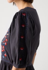 Dunkles Baumwollkleid mit Puffärmeln. Verfügt über farbenfrohe Blumenstickerei in Rot, Blau und Weiß entlang der Ärmel und des Oberteils.