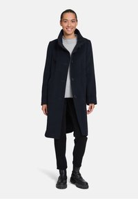 Marineblauwe wolmixjas met een hoge kraag, knoopsluiting en zijzakken; gedragen over een grijze top en zwarte broek, gecombineerd met zwarte laarzen.
