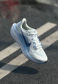 Bela mrežasta športna čevlja z svetlo modrim poudarkom in modrim logotipom swoosh, ki ima teksturirano površino in ukrivljen dizajn podplata.