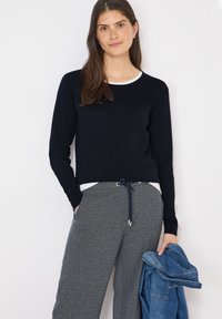 Femme aux longs cheveux bruns portant un pull noir et un pantalon gris à cordon, tenant une veste en jean bleu pliée dans la main droite.