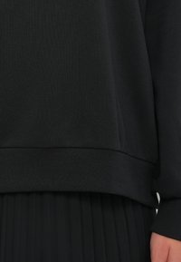 Svart sweatshirt med ribbede mansjetter og nederkant, med en glatt tekstur. Den er lagt over et plissert svart skjørt, noe som skaper et monokromatisk utseende.