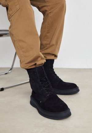Black suede ankle boots with lace-up design, textured surface, and rubber sole, accompanied by cuffed beige pants.