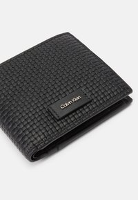 Must must valmistatud mustast kootud nahast, millel on sile tekstuur, ja sellel on metallist sildike "Calvin Klein". Kompaktne ristkülikukujuline disain.