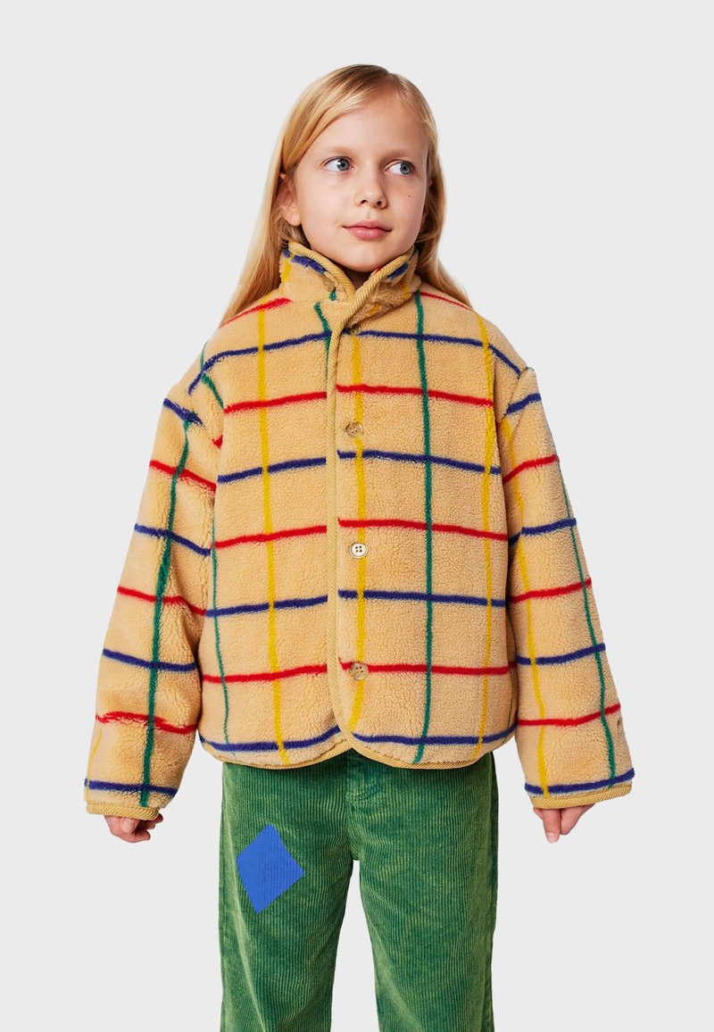 Giacca in pile beige con motivo a griglia colorata; progettata con un colletto alto, due bottoni e vestibilità comoda. Pantaloni in corduroy verde con una toppa blu.