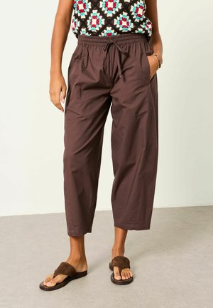 Person wearing loose brown cropped pants with drawstring waist, one hand in pocket, multicolored diamond-patterned top, and brown sandals.