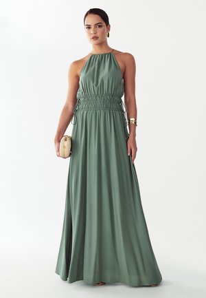 Kobieta ubrana w bezrękawową, szałwiowozieloną sukienkę maxi na szyję, trzymająca małą beżową kopertówkę, uzupełniona złotą biżuterią.