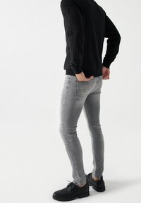 Personne portant un jean skinny gris, un pull noir et des chaussures noires à lacets, debout devant un fond clair uni.