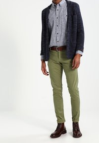 Mörkblå stickad cardigan över en ljusgrå skjorta med knappar, olivgröna byxor och bruna snörstövlar. Enkel, modern och texturerad design.