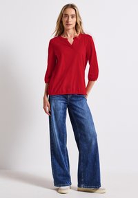 Rotes, langärmliges Oberteil mit Knopfausschnitt in V-Form und elastischen Bündchen, kombiniert mit lockeren, hellblauen Jeans im weiten Schnitt.