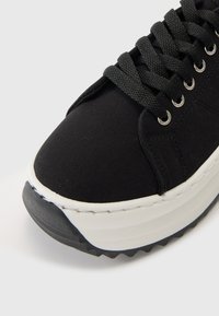 Svarte sneakers laget av lerret med en tykk hvit såle og teksturert svart gummiyttersåle. Har svarte snøringer og metalløyer.