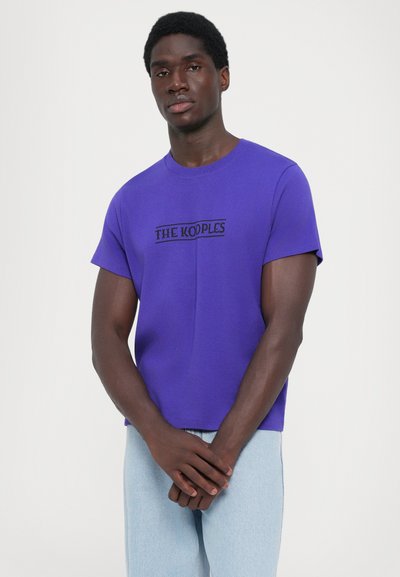 Μωβ κοντομάνικο μπλουζάκι από βαμβάκι, με μαύρο λογότυπο "THE KOOPLES" εμπρός, φορεμένο με ανοιχτό μπλε τζιν παντελόνι.