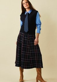 Stickad väst i marinblått över en ljusblå skjorta, tillsammans med en rutig kjol i mörka toner. Bruna knähöga stövlar fulländar looken.