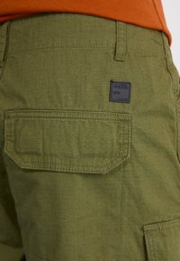 Olive zelene kratke hlače iz teksturiranega blaga, z ravnim zadnjim žepom s podrobnostmi šivanja in črno nalepko z blagovno znamko "ALPHA INDUSTRIES."
