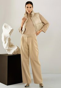 Vrouw in een beige coltrui, gewatteerde vest, wijde broek en slangenhuid laarzen staat naast een wit abstract beeldhouwwerk op een zwart voetstuk.