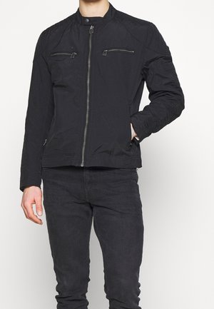 Mann trägt eine schwarze Jacke mit Reißverschluss und Brusttaschen mit Reißverschluss sowie schwarze Hose, steht mit der Hand in der Jackentasche vor weißem Hintergrund.