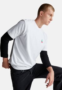 Mladý muž se naklání dopředu s jedním kolenem zvednutým, má na sobě bílé tričko s krátkým rukávem přes černé dlouhé rukávy a tmavé džíny.