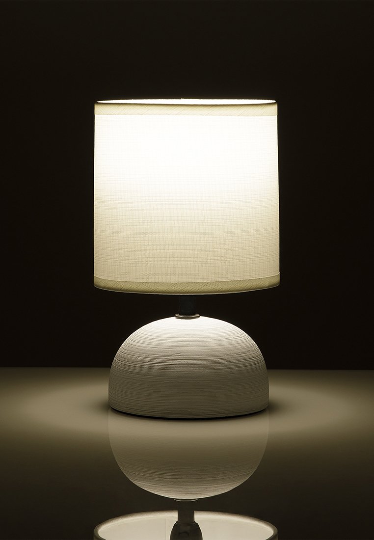 Stolní lampa s texturovaným kulatým bílým podstavcem a válcovým bílým textilním stínidlem, rozsvícená a vrhající měkké odrazy na povrch.