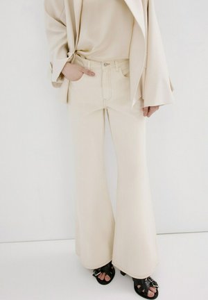 Persona vistiendo pantalones anchos beige, blazer y top a juego beige, con sandalias negras de tacón y puntera abierta sobre fondo blanco.