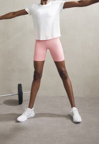 Camisa blanca de manga corta, pantalones cortos de ciclismo rosas y zapatillas blancas. El modelo está de pie con los brazos extendidos, mostrando el ajuste y diseño de la ropa.